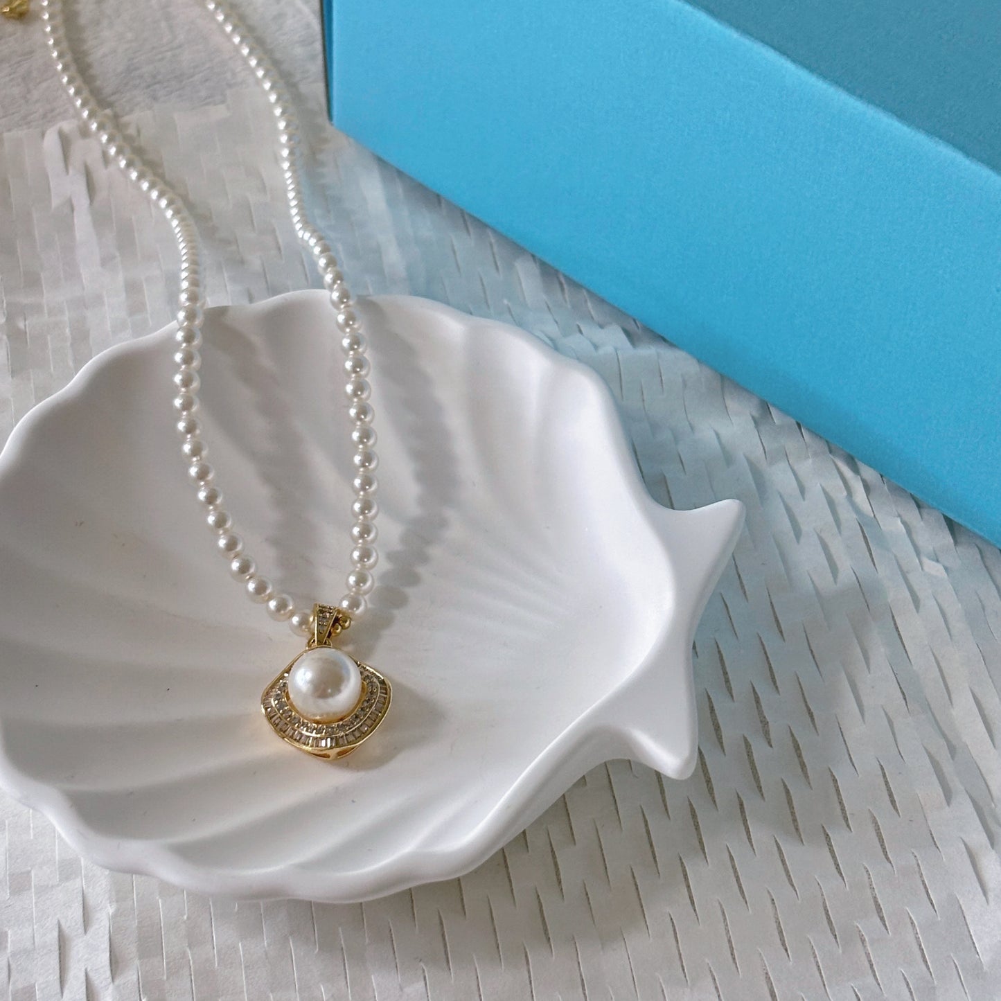 Pearl necklace with a gold pendant on a white shell, blue box in the background